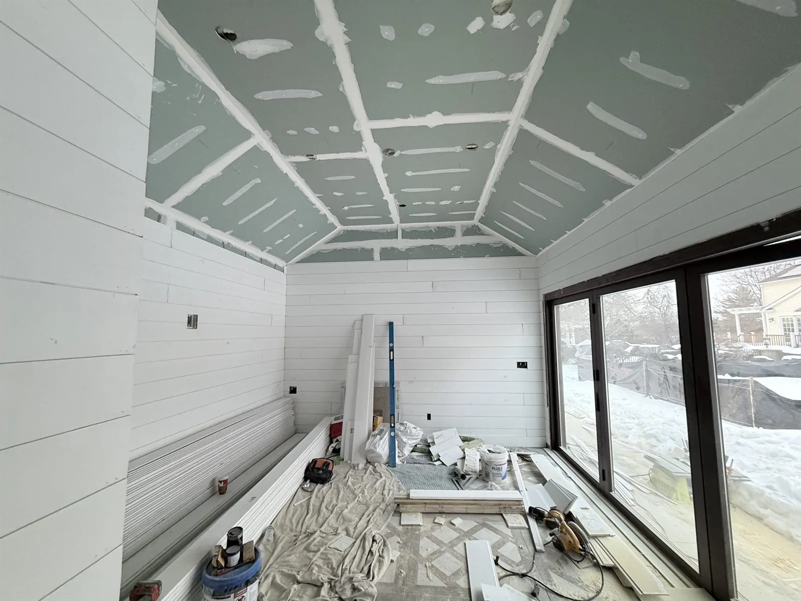 Sunroom under construction with unfinished drywall ceiling, taped seams, primed shiplap walls and tools on the floor